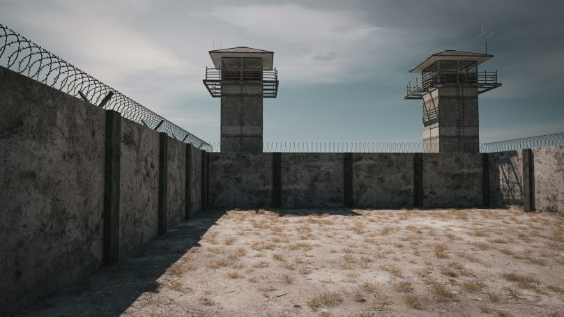 Custodial Facilities Concrete prison walls with two guard towers and a sandy courtyard under a cloudy sky.
